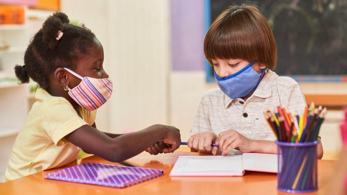 Zwei Kinder sitzen mit Mundschutz wegen Coronavirus-Pandemie an einem Tisch in der Grundschule oder Vorschule