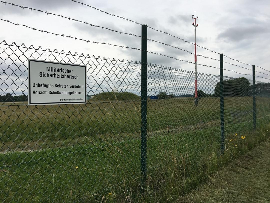 An einem hohen Zaun hängt ein Warnschild, darauf steht: "Militärischer Sicherheitsbereich. Unbefugtes Betreten verboten! Vorsicht Schußwaffengebrauch!"