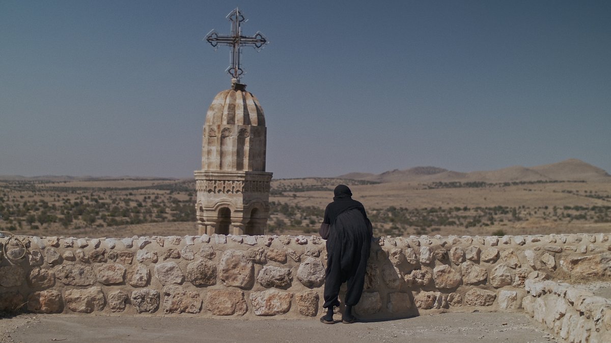 Schwarz gekleidete Frau schaut über eine Sandsteinmauer auf karges Land, vor ihr ein Kirchturm mit Kreuz.