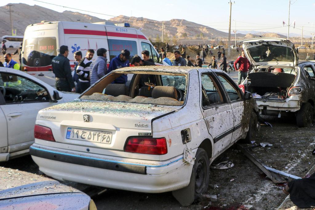 03.01.2024, Iran, Kerman: Menschen stehen neben zerstörten Autos nach einer Explosion.