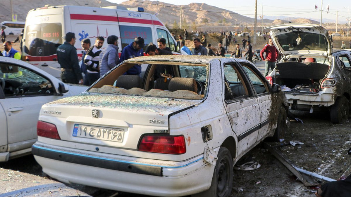 03.01.2024, Iran, Kerman: Menschen stehen neben zerstörten Autos nach einer Explosion.