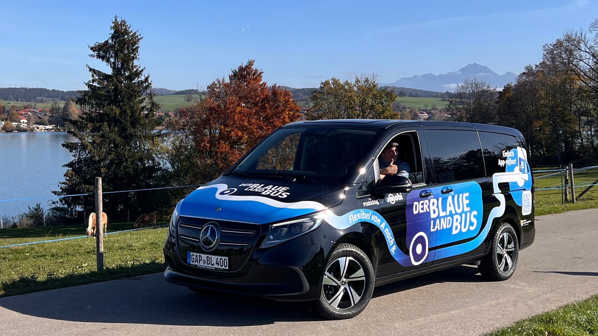 Ein Neunsitzer-Bus mit der Aufschrift "Der Blaue Land Bus" steht auf einer schmalen Straße vor einer Landschaft in Oberbayern; im Hintergrund eine Baumreihe, dahinter ein See und Berge.