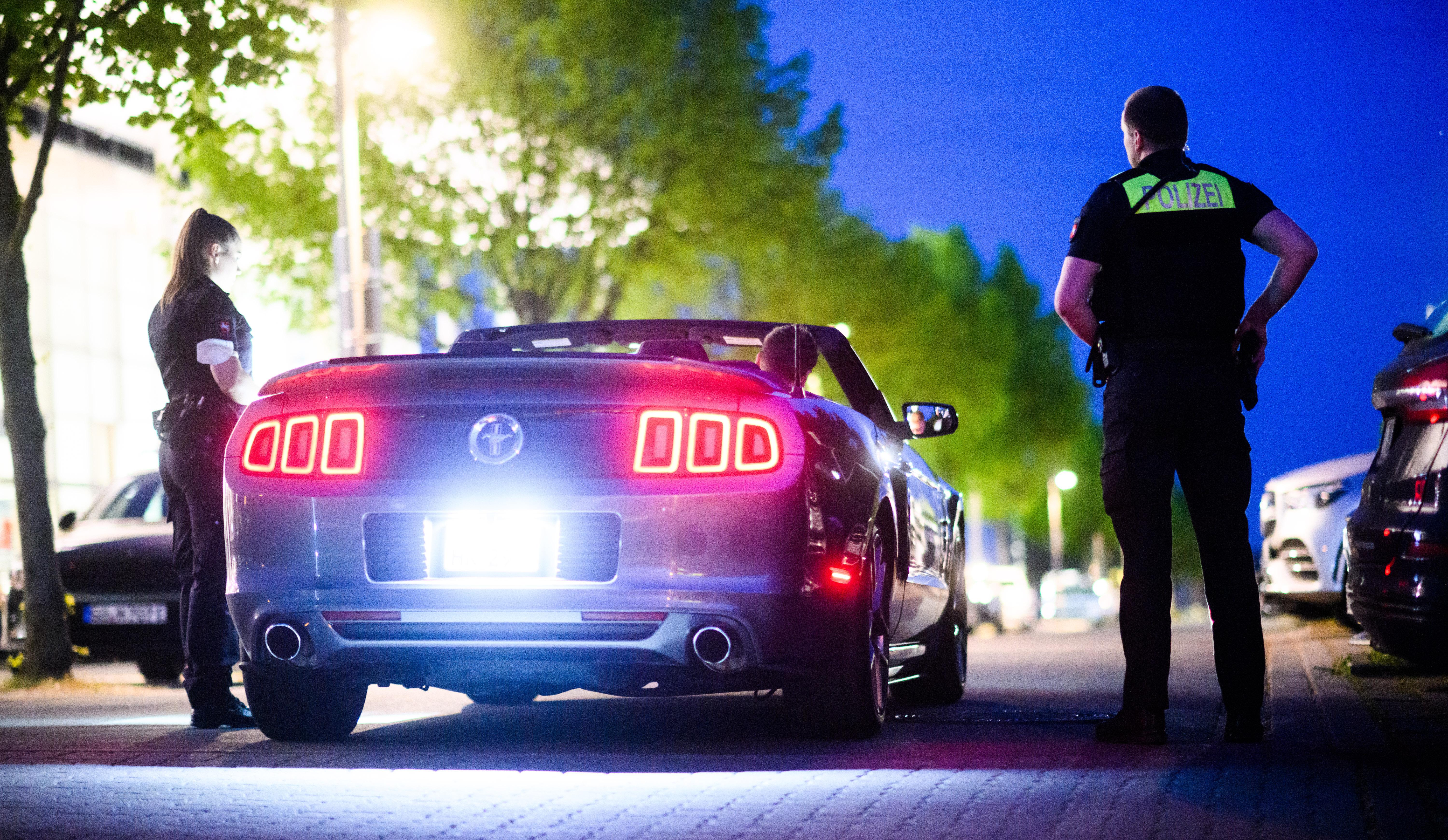 Eine Polizistin und ein Polizist stehen im Halbdunkel neben einem Luxus-Cabriolet und kontrollieren die Person am Steuer. (Symbol-/Archivbild)