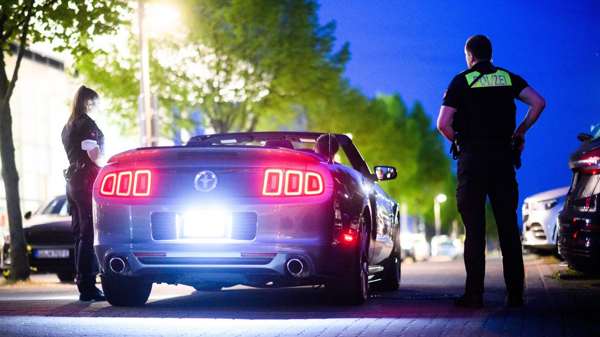 Eine Polizistin und ein Polizist stehen im Halbdunkel neben einem Luxus-Cabriolet und kontrollieren die Person am Steuer. (Symbol-/Archivbild)