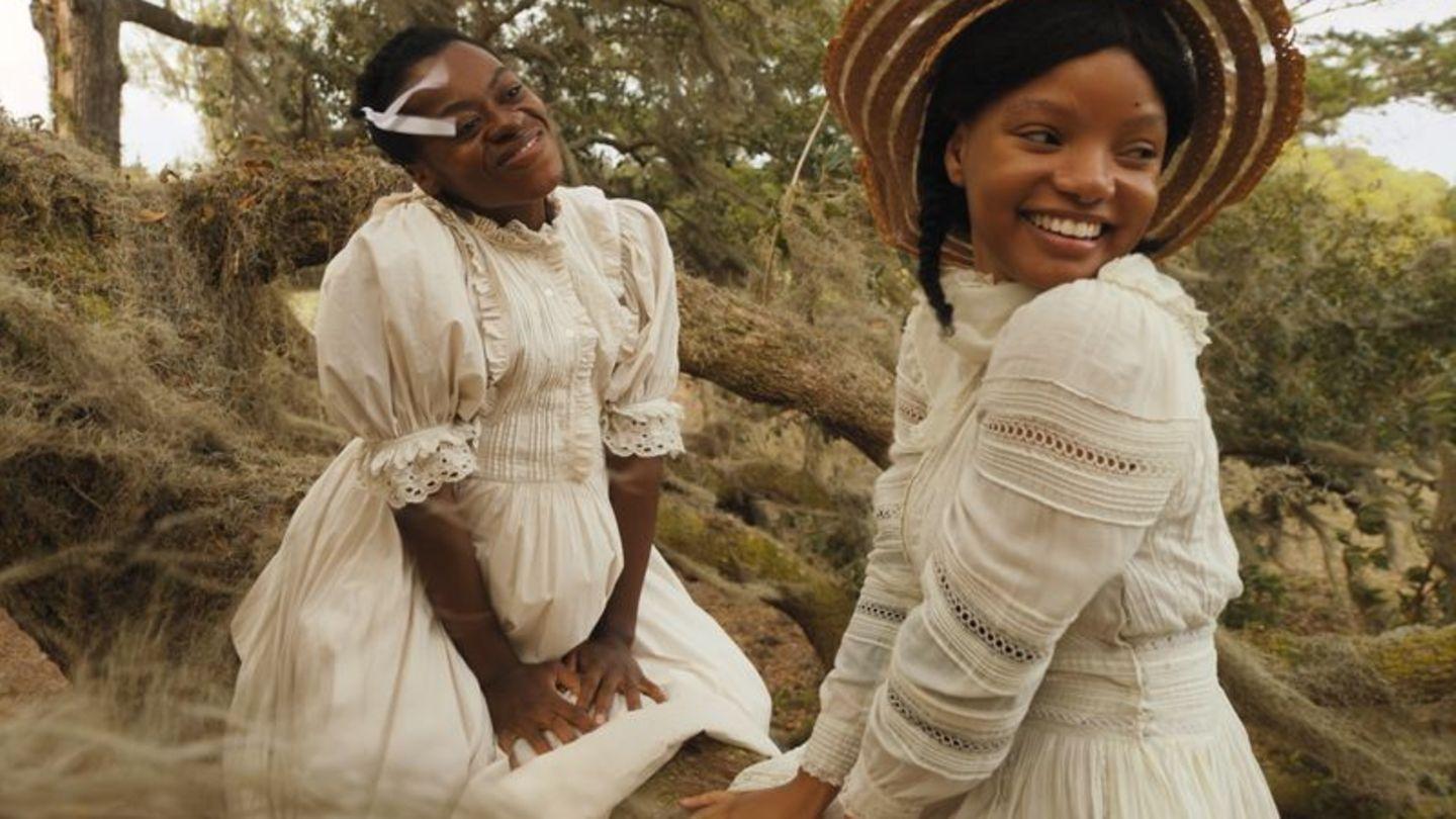 Zwei schwarze Frauen in altmodischer, weißer Kleidung lachen im Freien.  