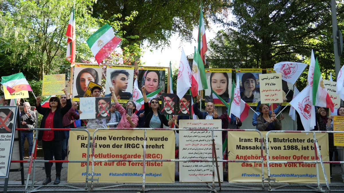 Hinter einer Absperrung schwenken iranische Flaggen und weisen mit Plakaten auf die Vergangenheit des verunglückten Präsidenten Raisi als Richter hin.