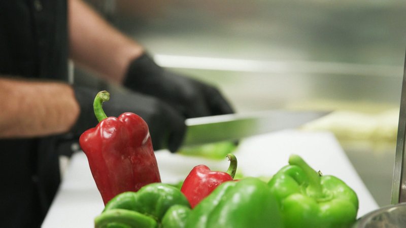 Ein Koch schneidet mit schwarzen Handschuhen rote und grüne Paprika.