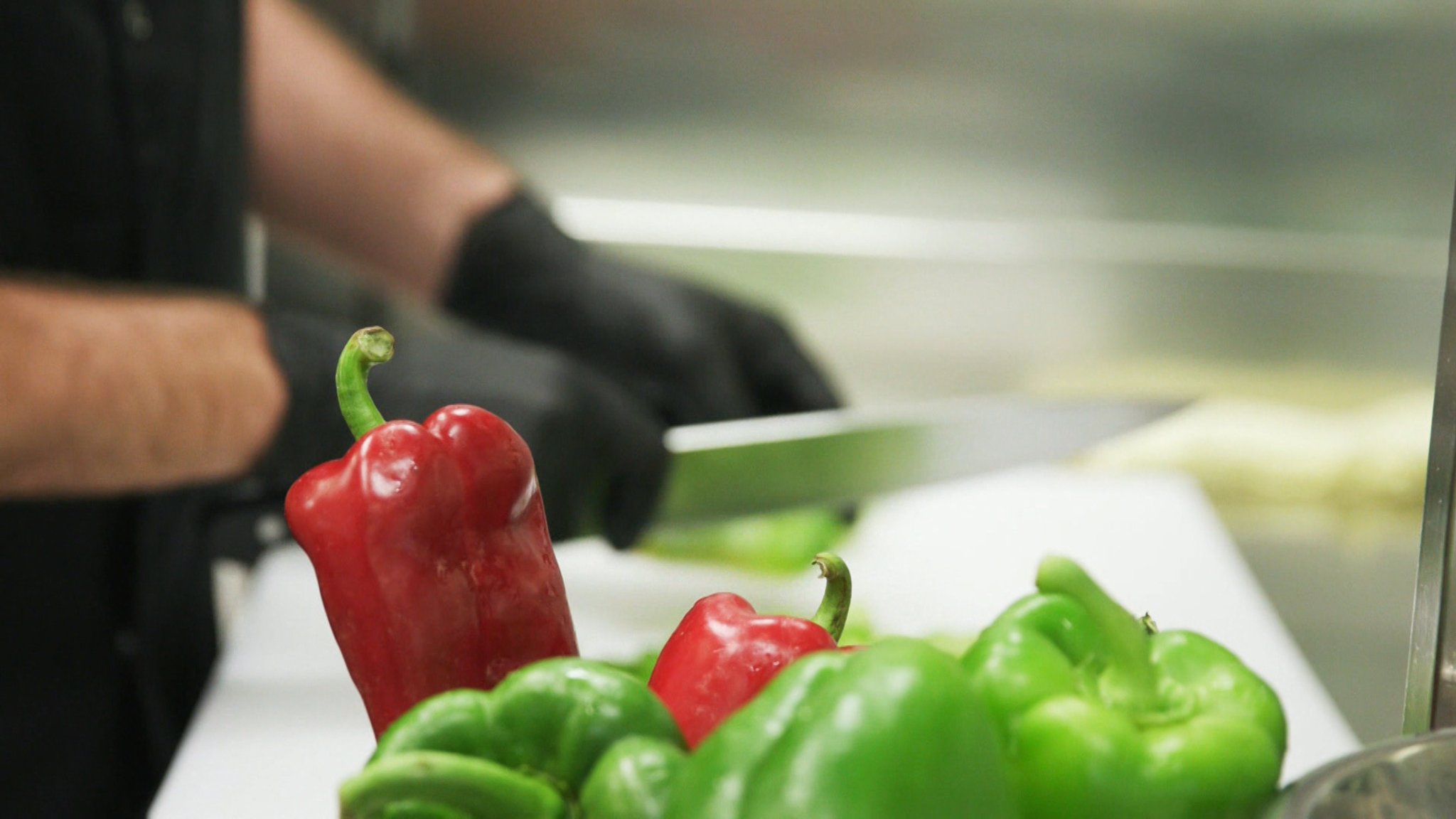 Ein Koch schneidet mit schwarzen Handschuhen rote und grüne Paprika.