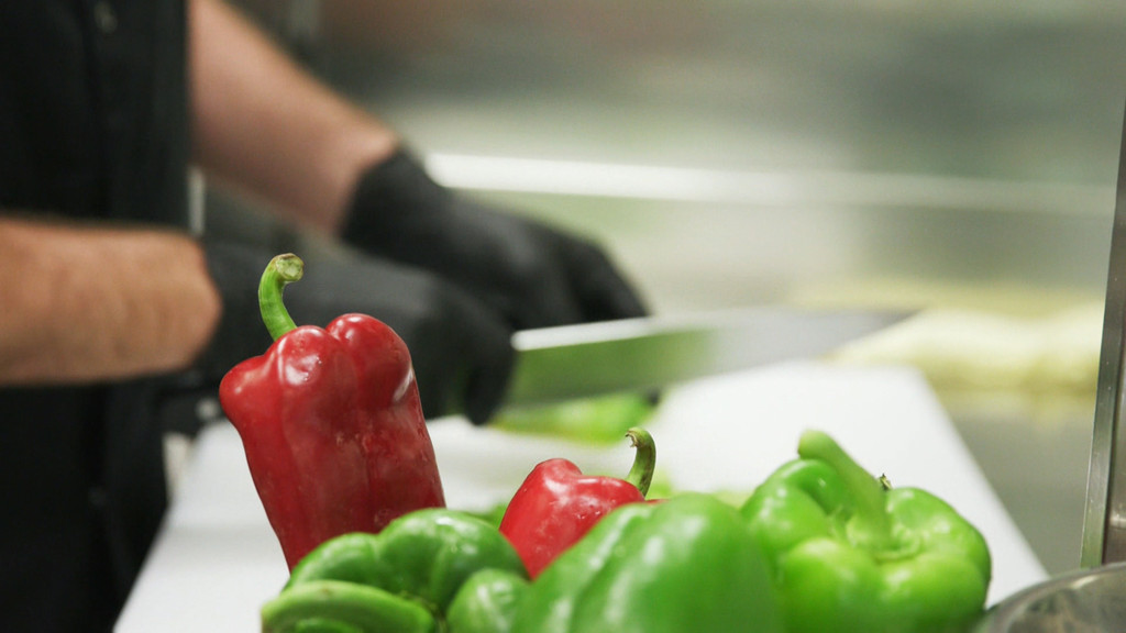 Ein Koch schneidet mit schwarzen Handschuhen rote und grüne Paprika.