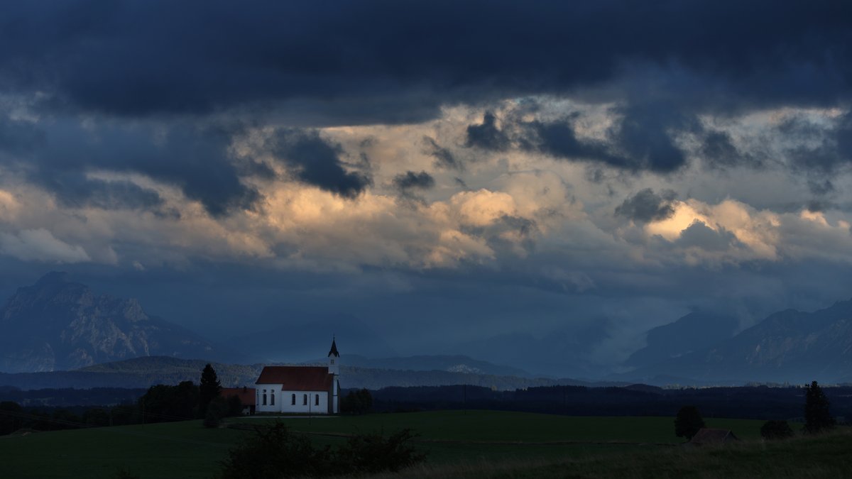 Die tief stehende Sonne beleuchtet einen Wolkenstreifen über der am Alpenrand gelegenen Wallfahrtskirche Sankt Alban.