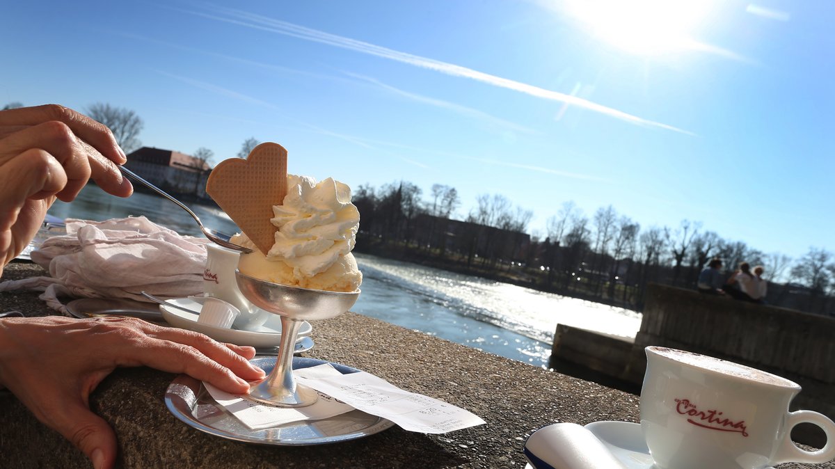 Ein Vanilleeis mit Sahne und zwei Tassen Kaffee stehen vor einer Eisdiele am Lech auf einer Mauer.