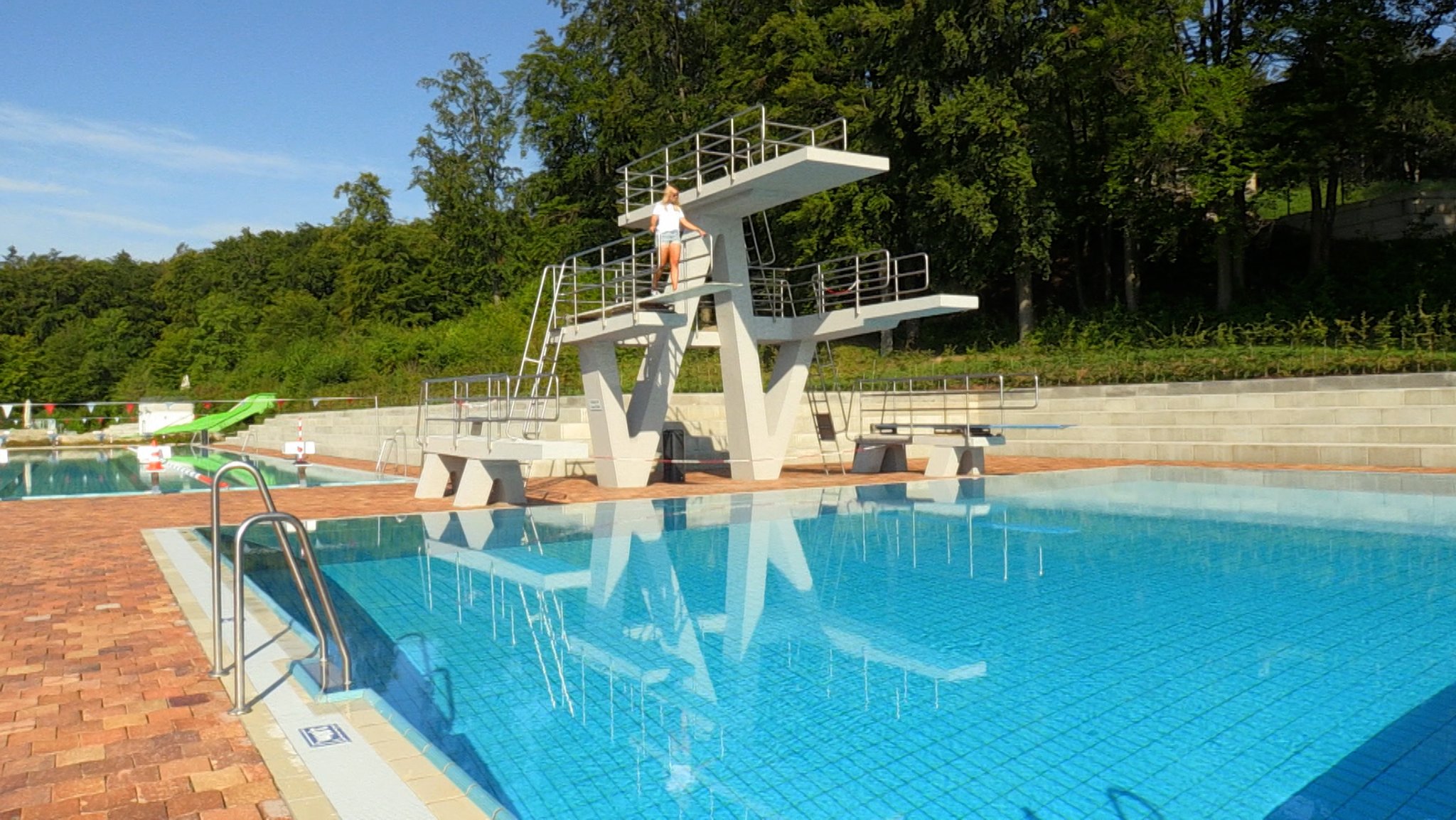 Eine Frau steht auf einem Drei-Meter-Brett in einem Freibad. Das Becken ist leer und es sind keine Menschen da.