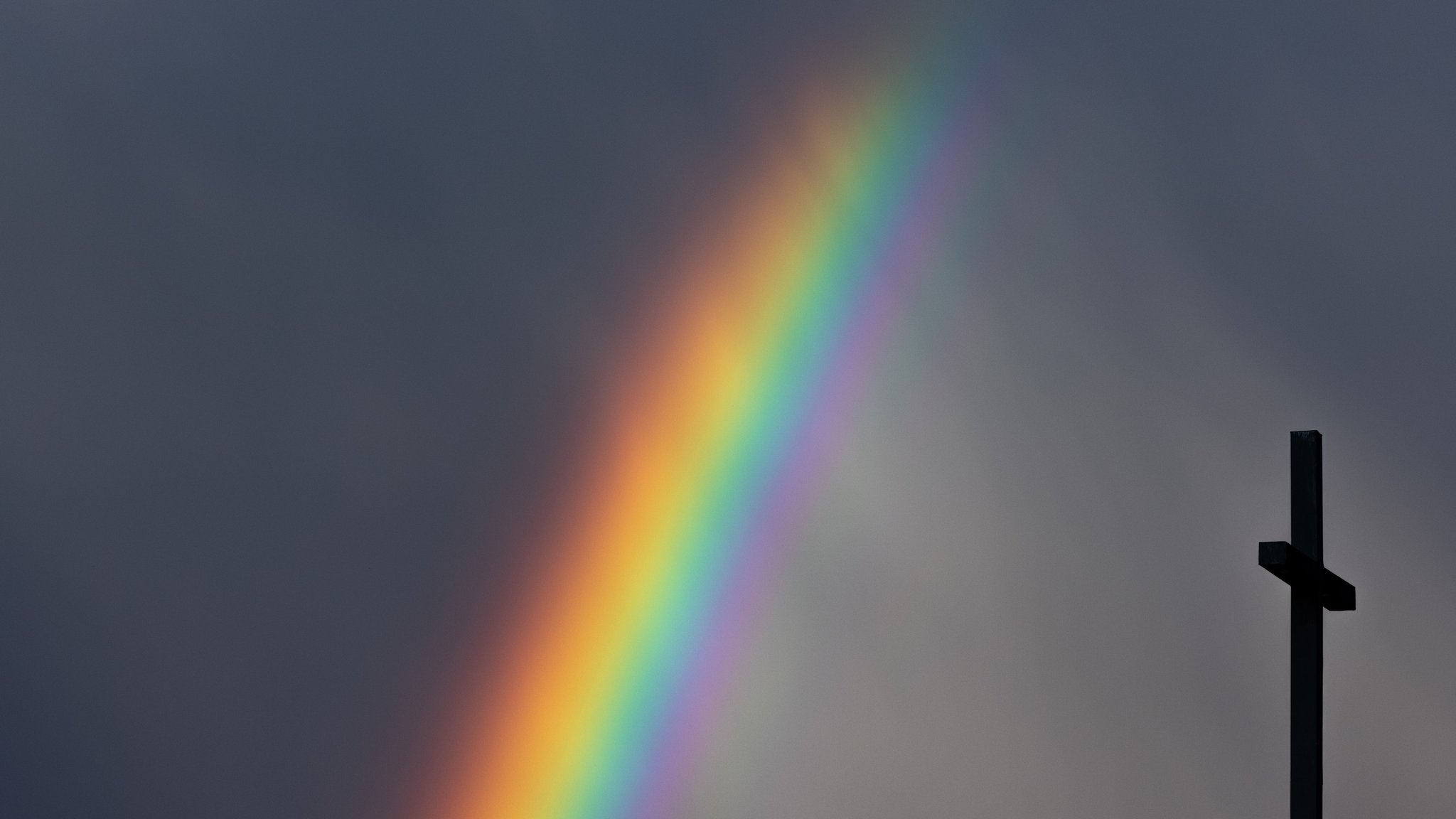 Ein kräftiger Regenbogen ist am dunklen Himmel neben dem Kreuz einer Kirche zu sehen.