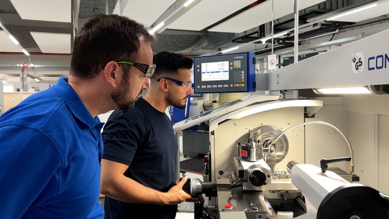 Ausbilder Dodik und Azubi Yasseen stehen an der Drehmaschine und fertigen ein Bauteil als Vorbereitung auf die Zwischenprüfung der Ausbildung zur Fachkraft Metalltechnik.