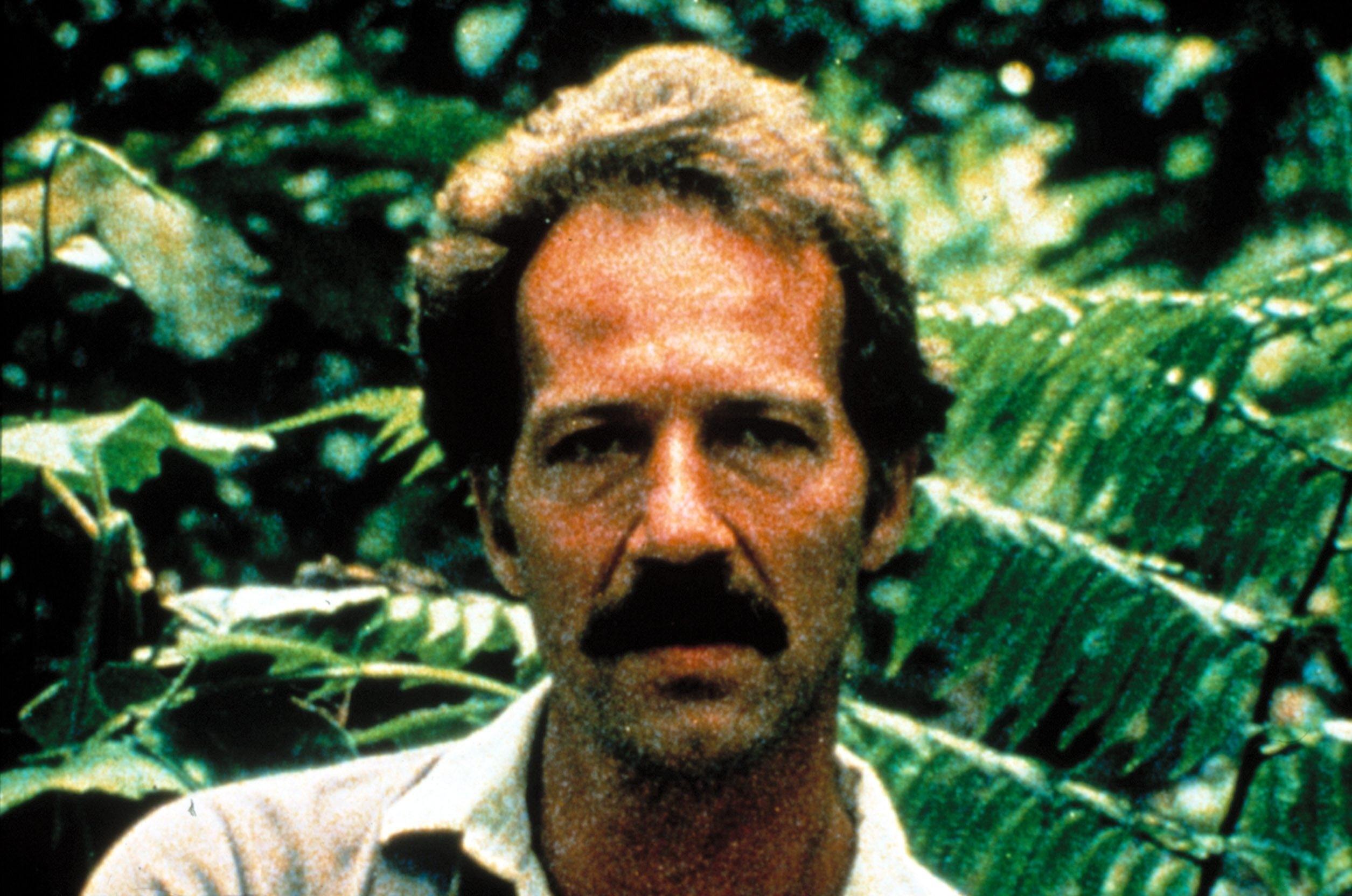  Porträt-Foto von Werner Herzog mit Schnurrbart vor Urwaldhintergrund.