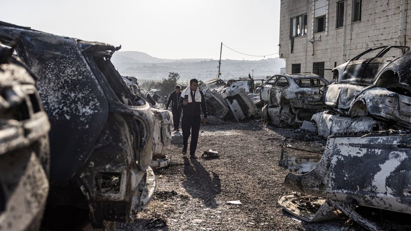 27.02.2023, Palästinensische Gebiete, Huwara: Männer gehen an verbrannten und zerstörten Fahrzeugen in der Stadt Huwara vorbei.