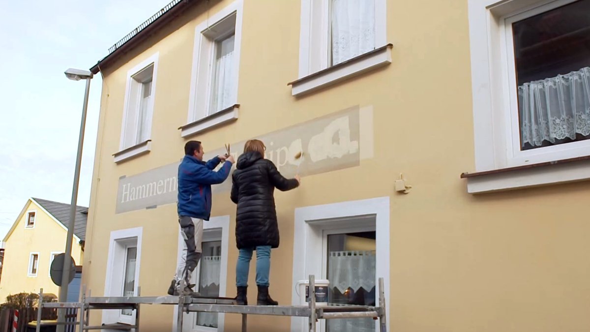 Ein Mann und eine Frau bringen an der Fassade eines Hauses einen Schriftzug an. 