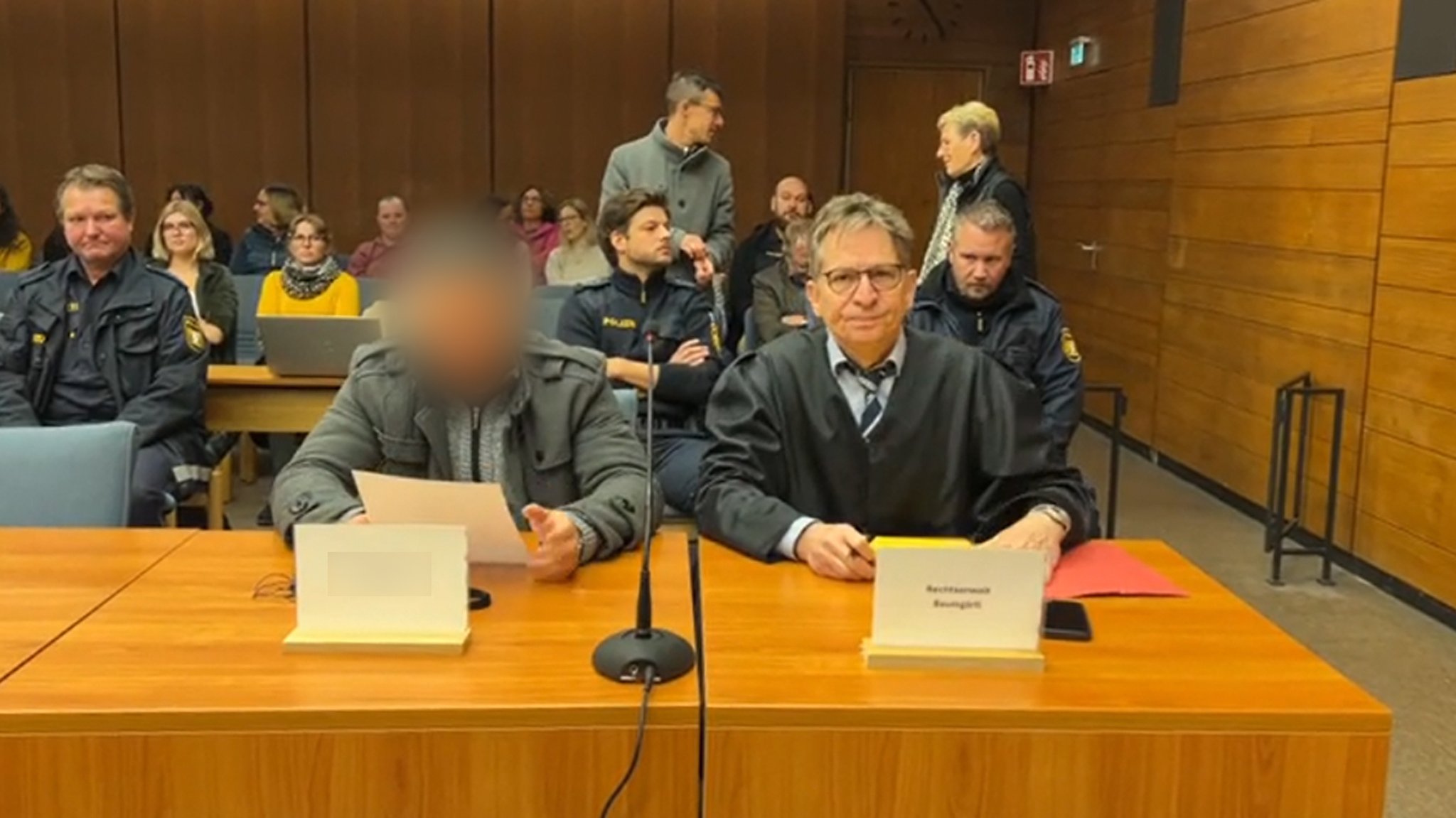 In einem Sitzungssaal des Gerichts sitzt der Hauptangeklagte neben seinem Verteidiger. Im Hintergrund sind Polizisten und Zusehende zu sehen. 