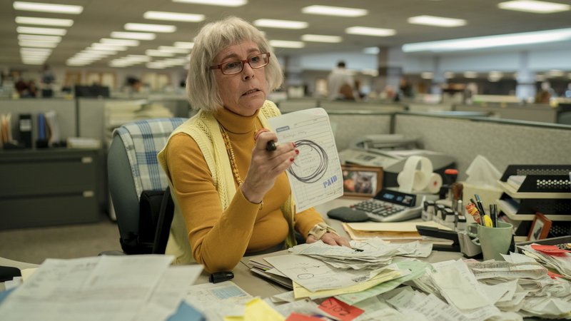 Filmbild aus "Everything Everywhere All At Once": Eine Frau sitz in gelben Pullover und Strickweste an einem Büroschreibtisch voller Papiere und hält ein Formular hoch
