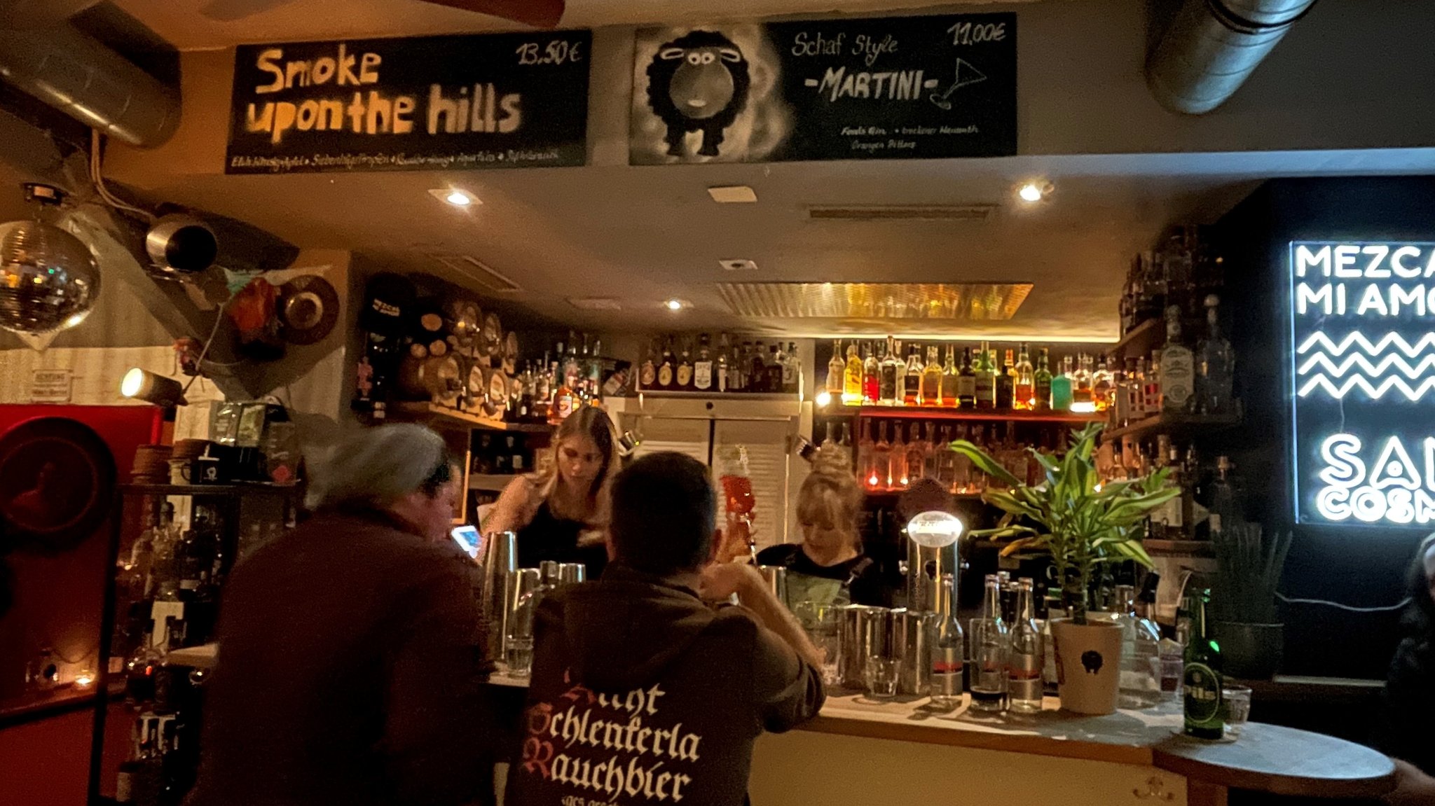 Zwei Menschen sitzen an einer Bar, dahinter stehen zwei Frauen.