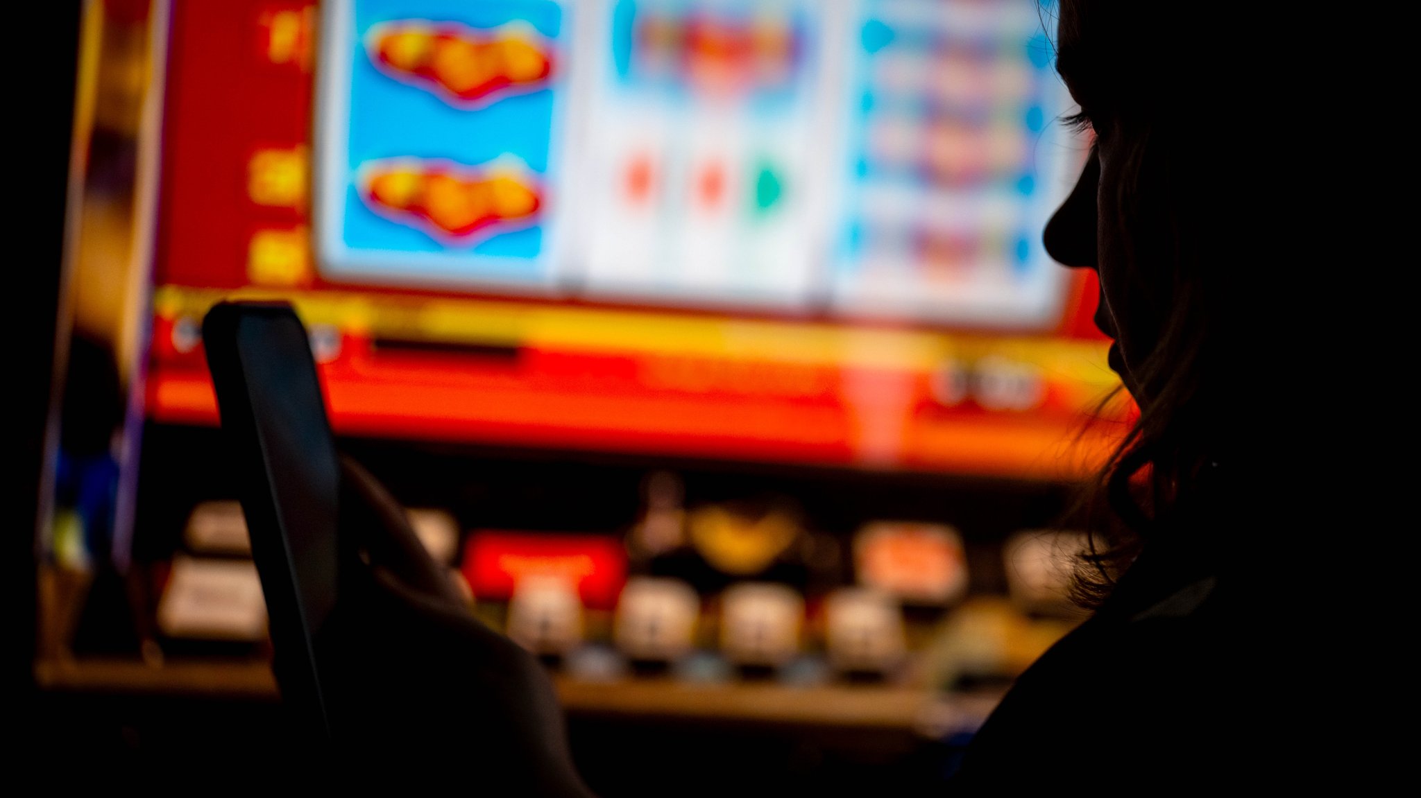 (Symbolbild) Eine Frau steht vor einem Spielautomaten und blickt auf ihr Smartphone