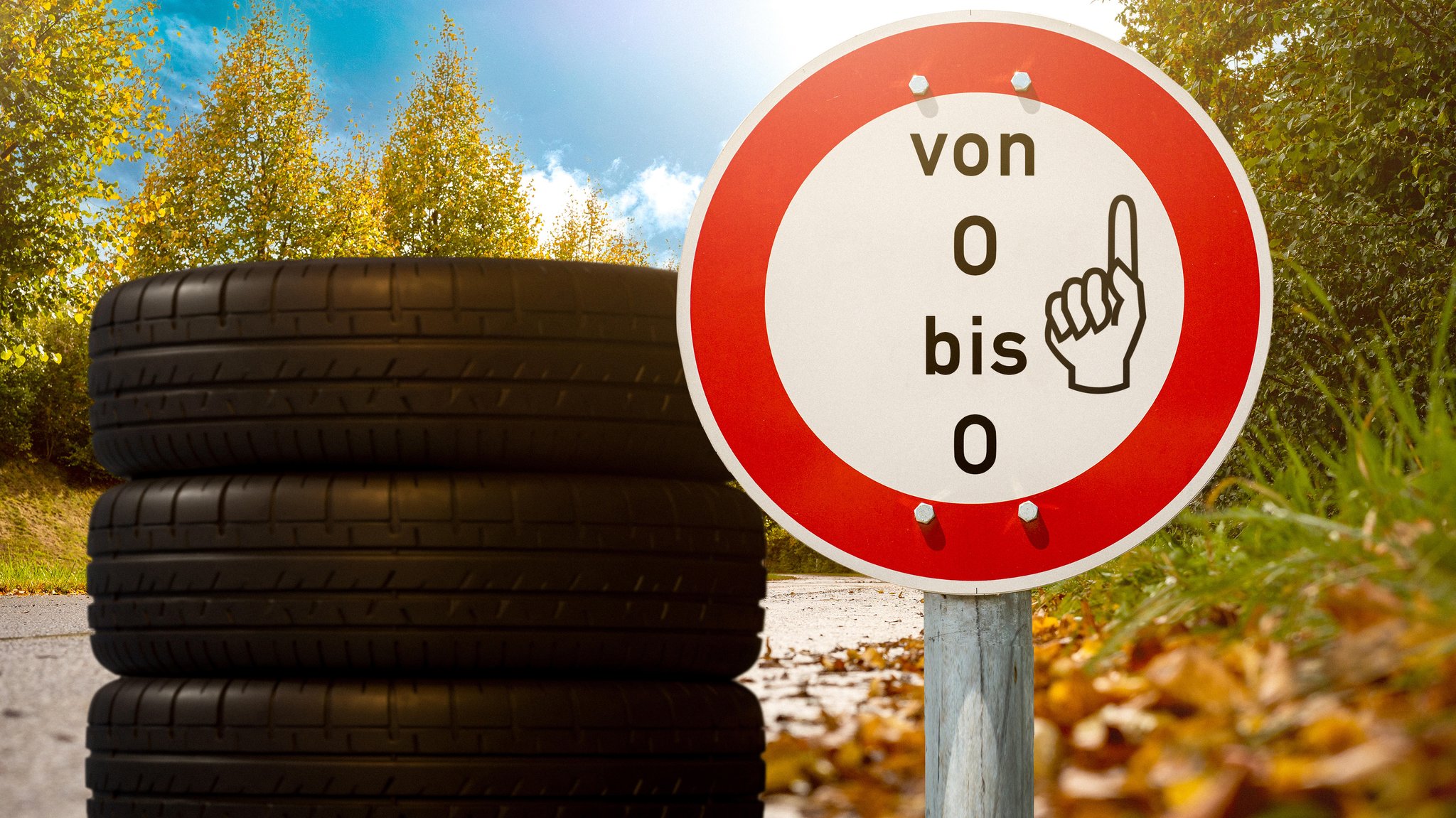 Ein Stapel Winterreifen vor herbstlicher Landschaft mit einem Schild mit der Aufschrift: Von O bis O