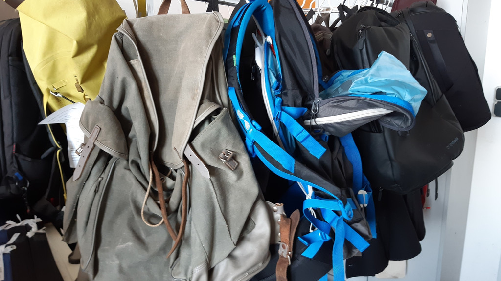 Ein alter grauer und ein neuer blauer Rucksack mit Reparaturbedarf hängen an Kleiderständer, dahinter schwarze Rucksäcke und Taschen und ein gelber Schutzsack   