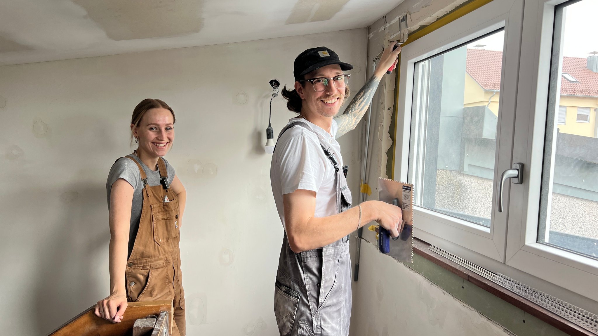 Max Fehr trägt Spachtelmasse an den Fensterkanten der Wand auf. Neben ihm steht Jule Günther und schaut genau hin.