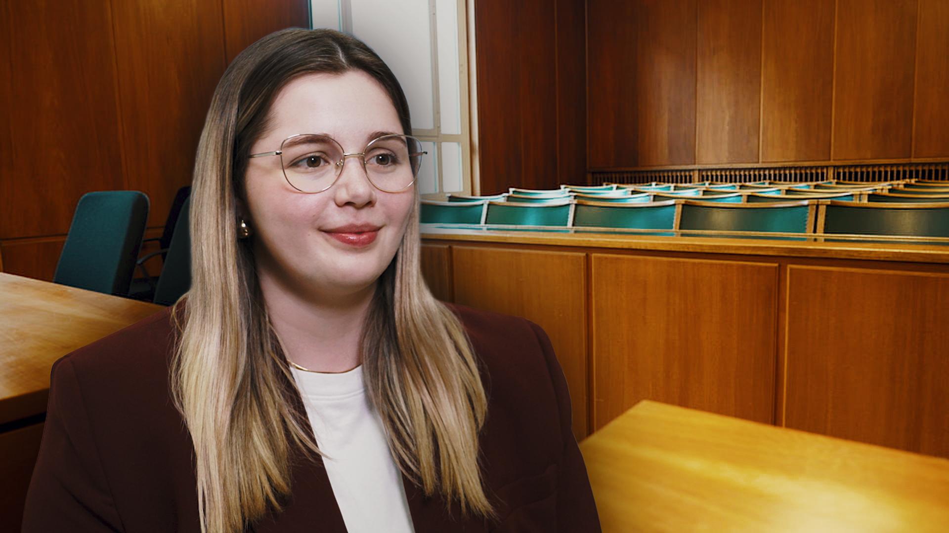 Im Vordergrund sitzt eine junge Frau mit Bille. Im Hintergrund zu sehen ist die Zuschauerbank eines Gerichtssaals.