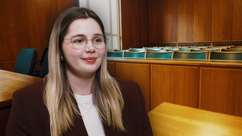 Im Vordergrund sitzt eine junge Frau mit Bille. Im Hintergrund zu sehen ist die Zuschauerbank eines Gerichtssaals. | Bild: Bayerischer Rundfunk 2026 Im Vordergrund sitzt eine junge Frau mit Bille. Im Hintergrund zu sehen ist die Zuschauerbank eines Gerichtssaals.