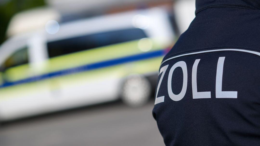 Zollbeamte stehen bei einer Kontrolle vor einem Polizeifahrzeug. 