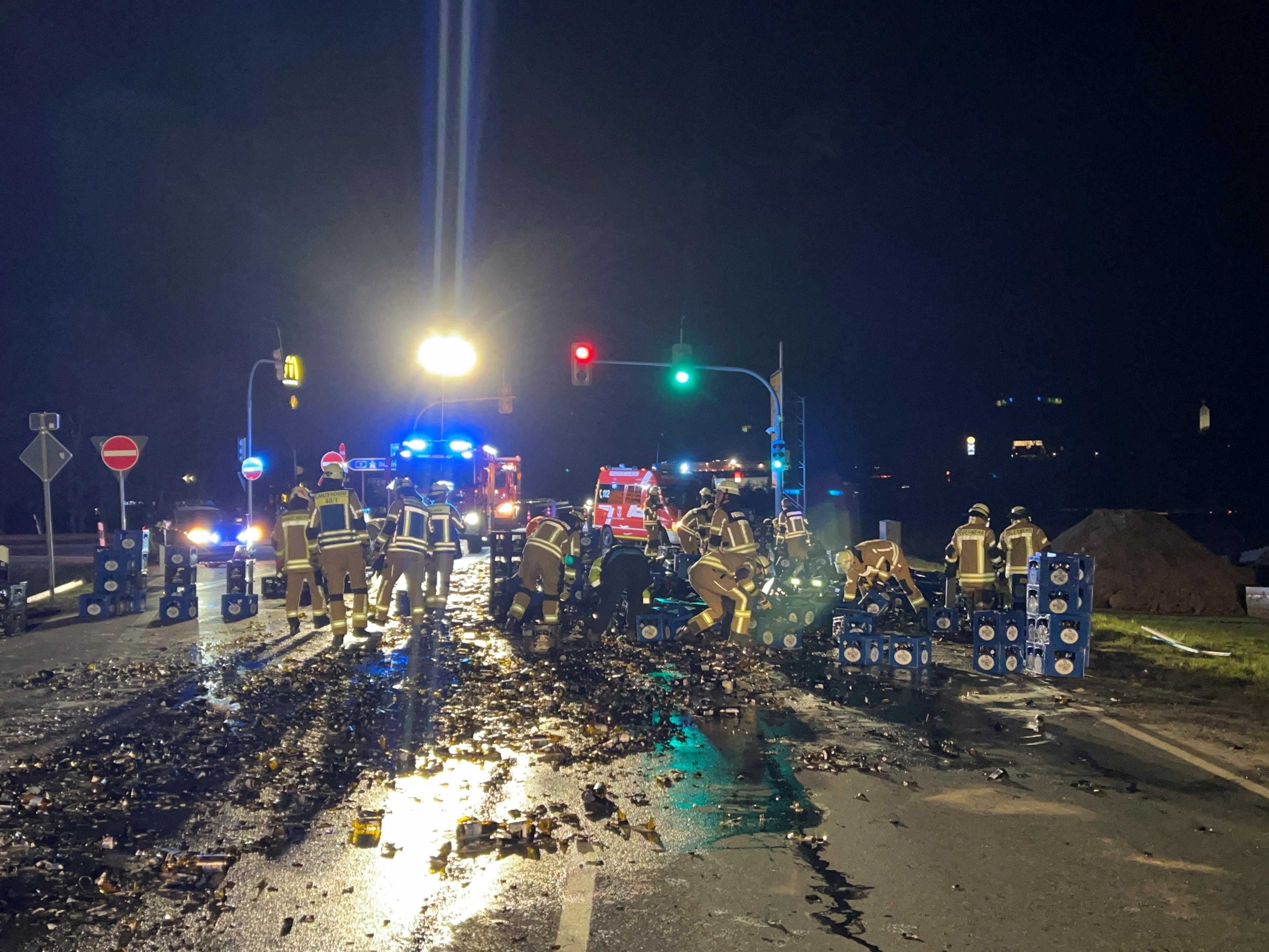 Eine Straße ist nachts mit zerbrochenen Bierflaschen übersäht, Feuerwehrleute räumen auf.