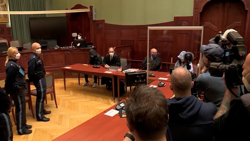 In einem Gerichtssaal filmen und fotografieren Kameraleute die Angeklagten, daneben stehen Polizeibeamte mit Mundschutz. 