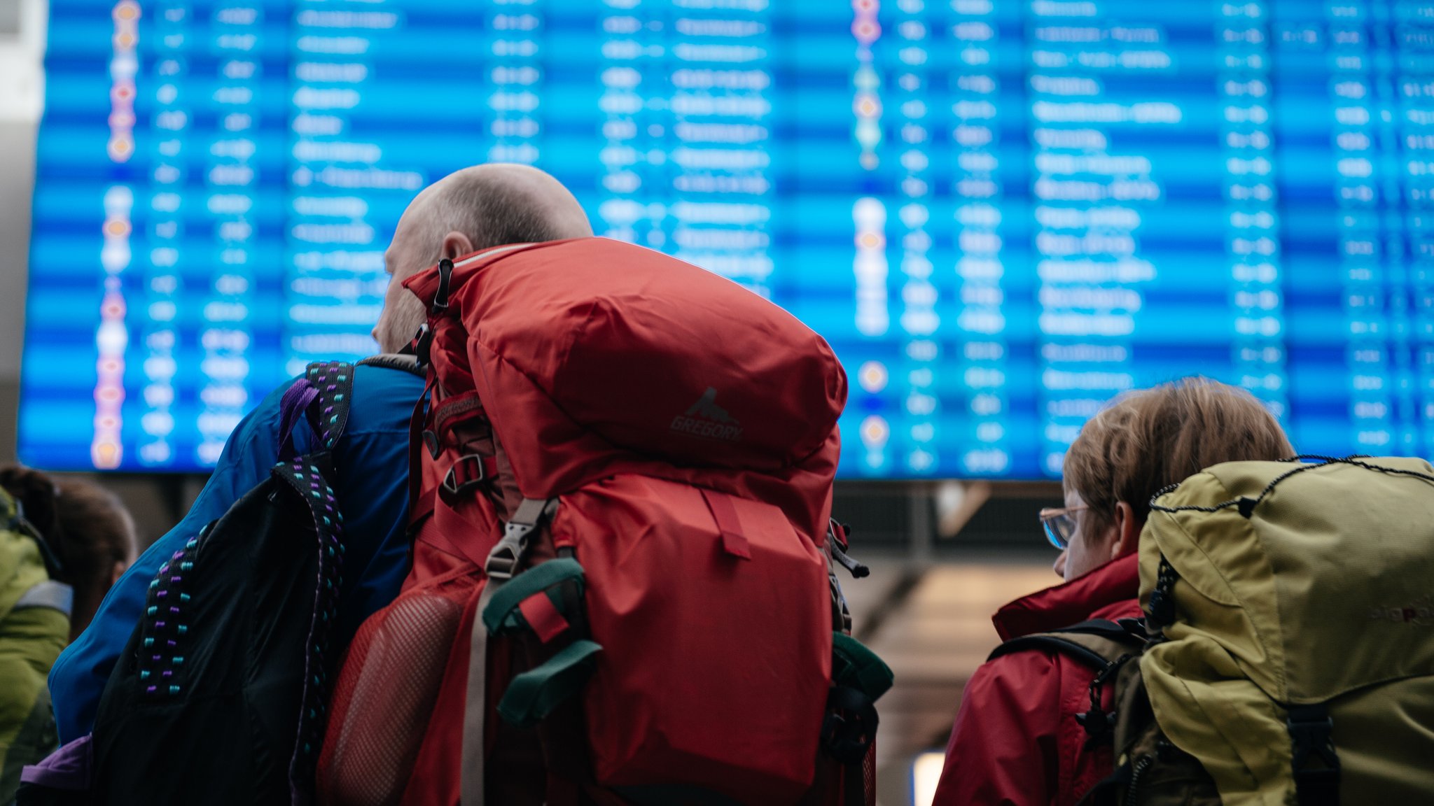 Zwei Menschen mit Rucksäcken warten vor einer Informationstafel am Flughafen