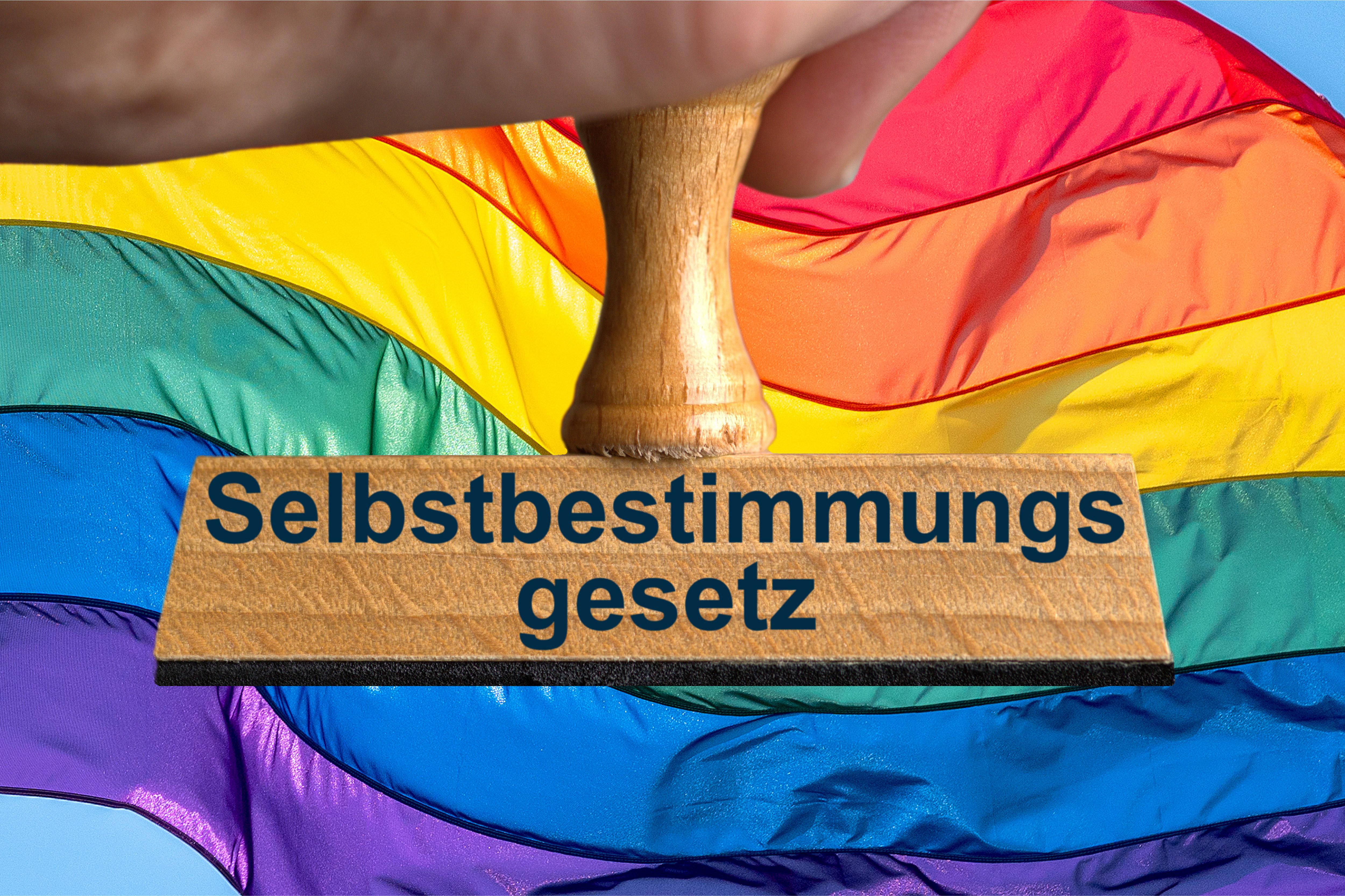 Ein Symbolbild, das einen Stempel mit der Aufschrift "Selbstbestimmungsgesetz" vor einer bunt gestreiften Flagge zeigt.