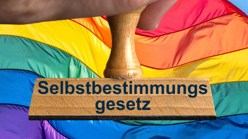 Ein Symbolbild, das einen Stempel mit der Aufschrift "Selbstbestimmungsgesetz" vor einer bunt gestreiften Flagge zeigt. | Bild: picture alliance / SULUPRESS.DE | Torsten Sukrow / SULUPRESS.DE Ein Symbolbild, das einen Stempel mit der Aufschrift "Selbstbestimmungsgesetz" vor einer bunt gestreiften Flagge zeigt.