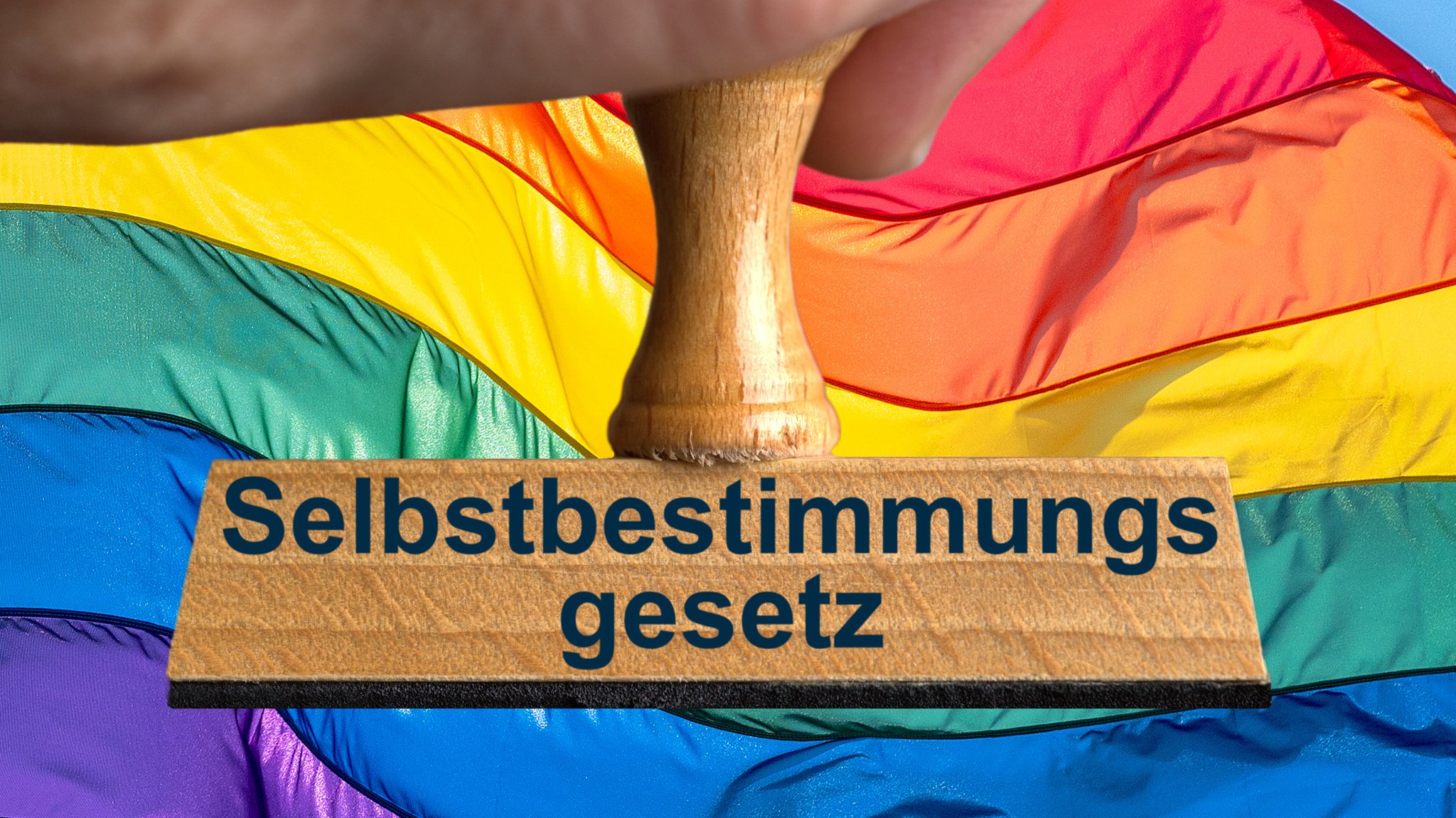 Ein Symbolbild, das einen Stempel mit der Aufschrift "Selbstbestimmungsgesetz" vor einer bunt gestreiften Flagge zeigt.