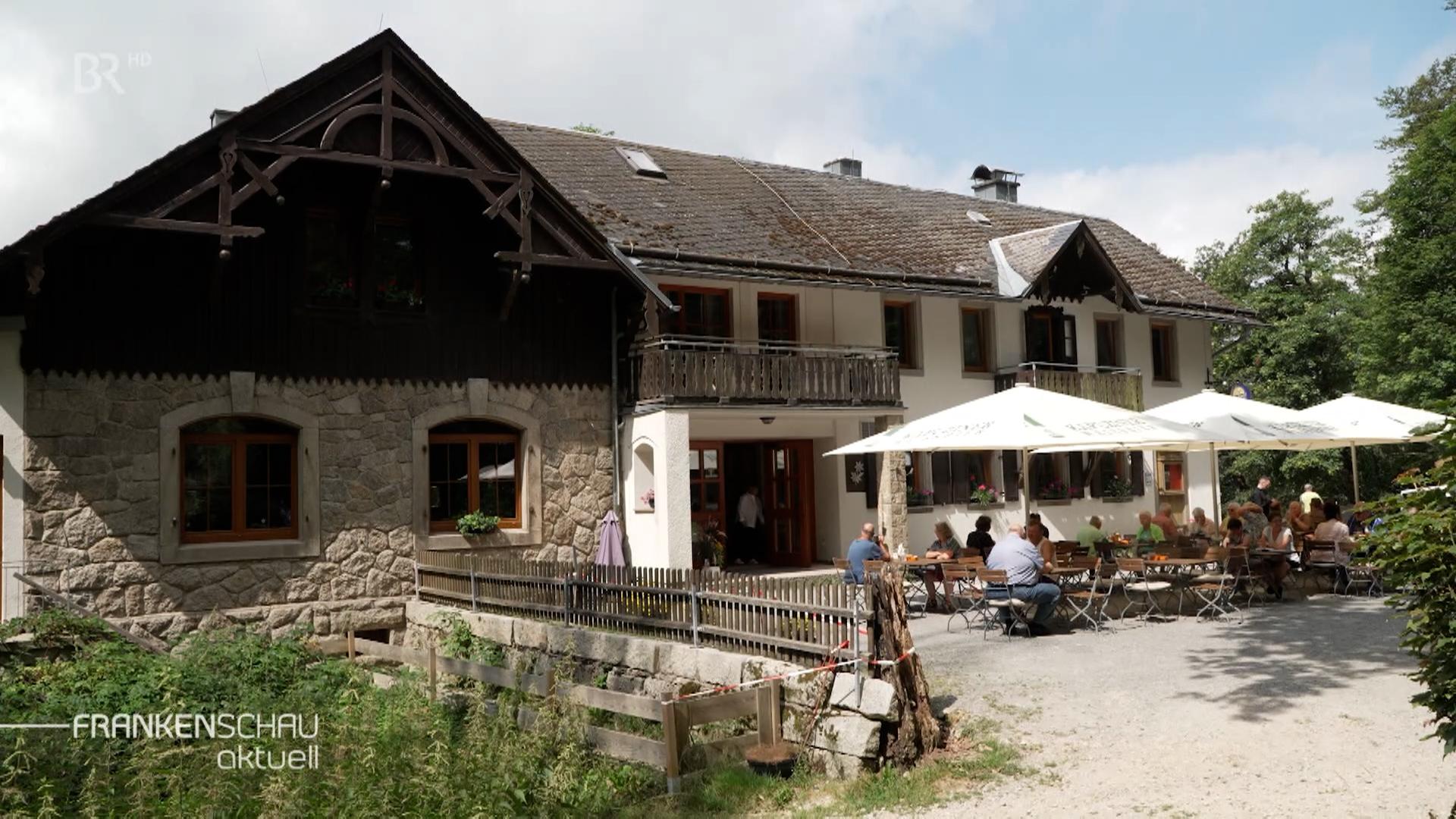 Vor einem historischen Gasthaus sitzen Besucher an Tischen unter Sonnenschirmen. 