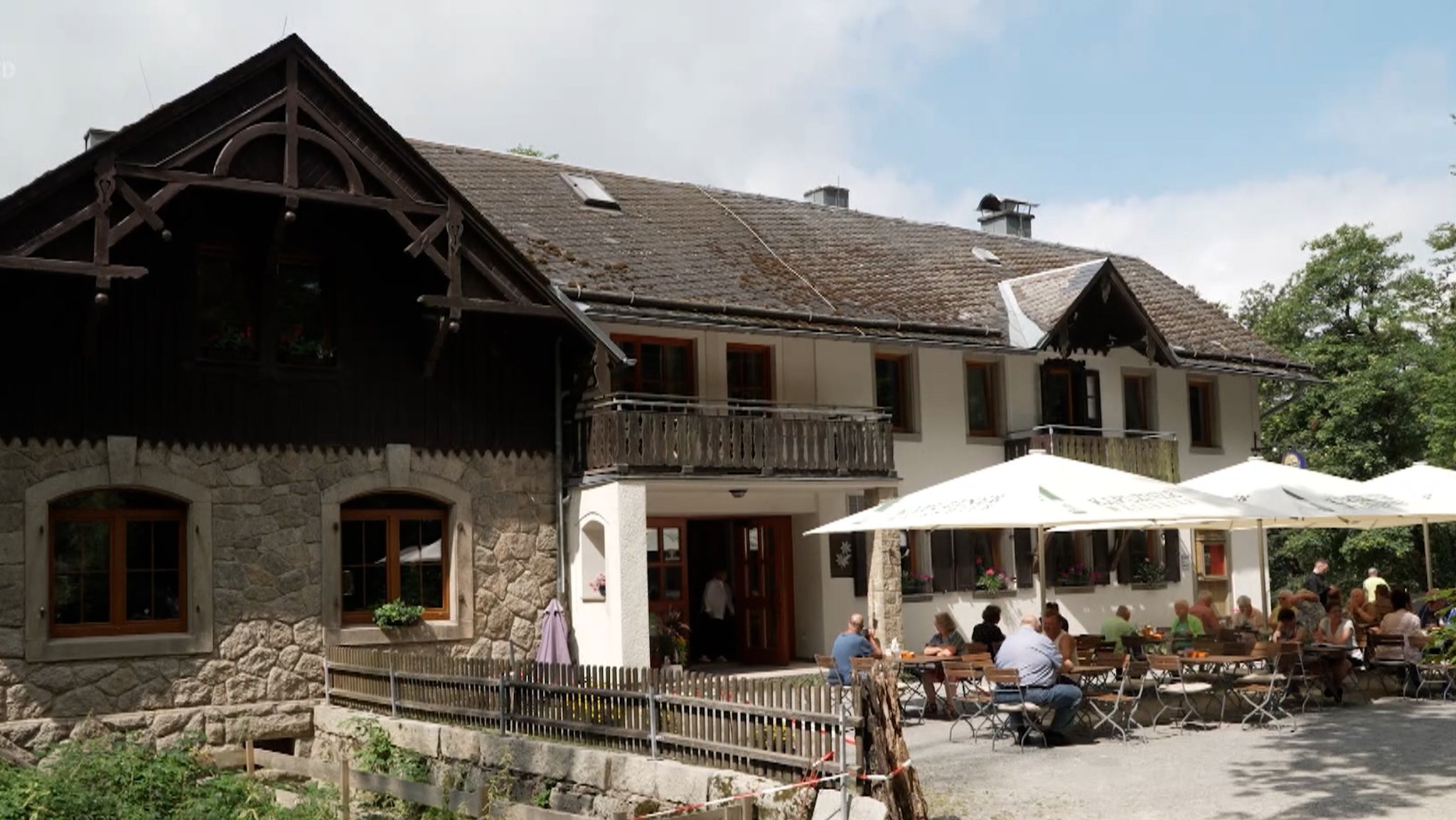 Vor einem historischen Gasthaus sitzen Besucher an Tischen unter Sonnenschirmen. 