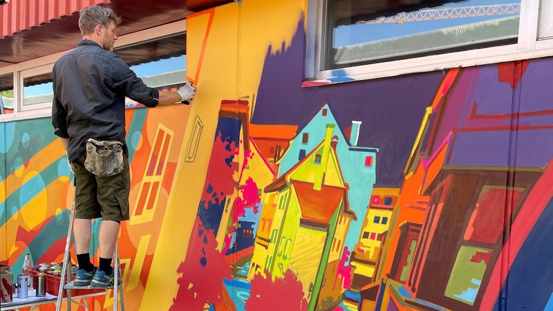 Künstler Daniel Döbner steht auf einer Leiter und sprüht mit einer Spraydose an die Wand die Haunstetter Hallenbades. Dort soll ein großes Kunstwerk rund um das Thema "Wasser" entstehen. | Bild: BR/Michaela Fembacher Künstler Daniel Döbner steht auf einer Leiter und sprüht mit einer Spraydose an die Wand die Haunstetter Hallenbades. Dort soll ein großes Kunstwerk rund um das Thema "Wasser" entstehen.