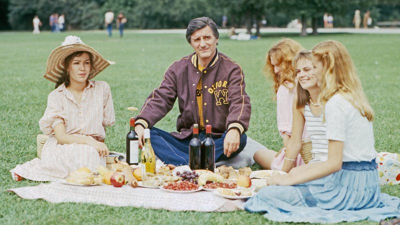 Helmut Fischer sitzt als "Monaco Franze" mit vier Schauspielerinnen auf einer Wiese. | Bild: picture alliance/United Archives | kpa Helmut Fischer sitzt als "Monaco Franze" mit vier Schauspielerinnen auf einer Wiese.