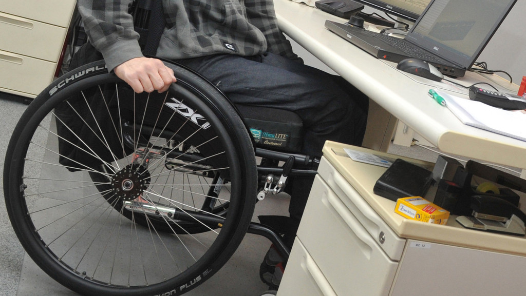 Ein Mann sitzt in einem Rollstuhl an einem Arbeitsplatz am Schreibtisch. 