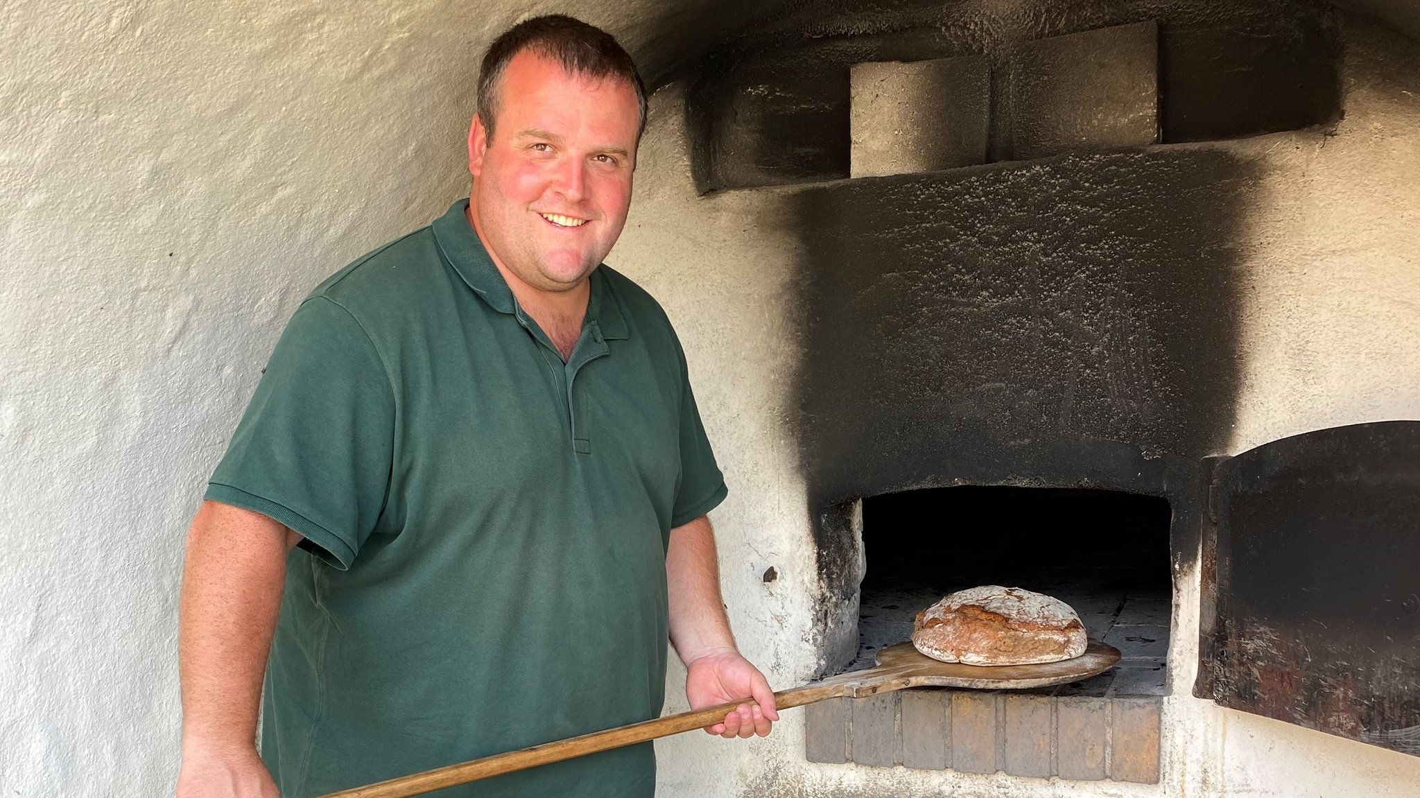 Ein Mann steht vor einem alten Backofen und hält auf einem Schieber ein Brot. 