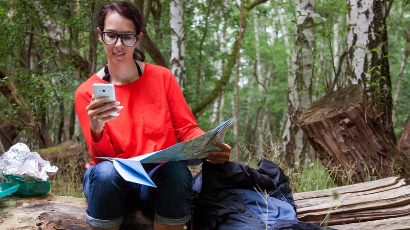 Frau macht Rast auf einem Baumstamm mit Handy und Karte in der Hand. | Bild: picture alliance / Christin Klose Frau macht Rast auf einem Baumstamm mit Handy und Karte in der Hand.