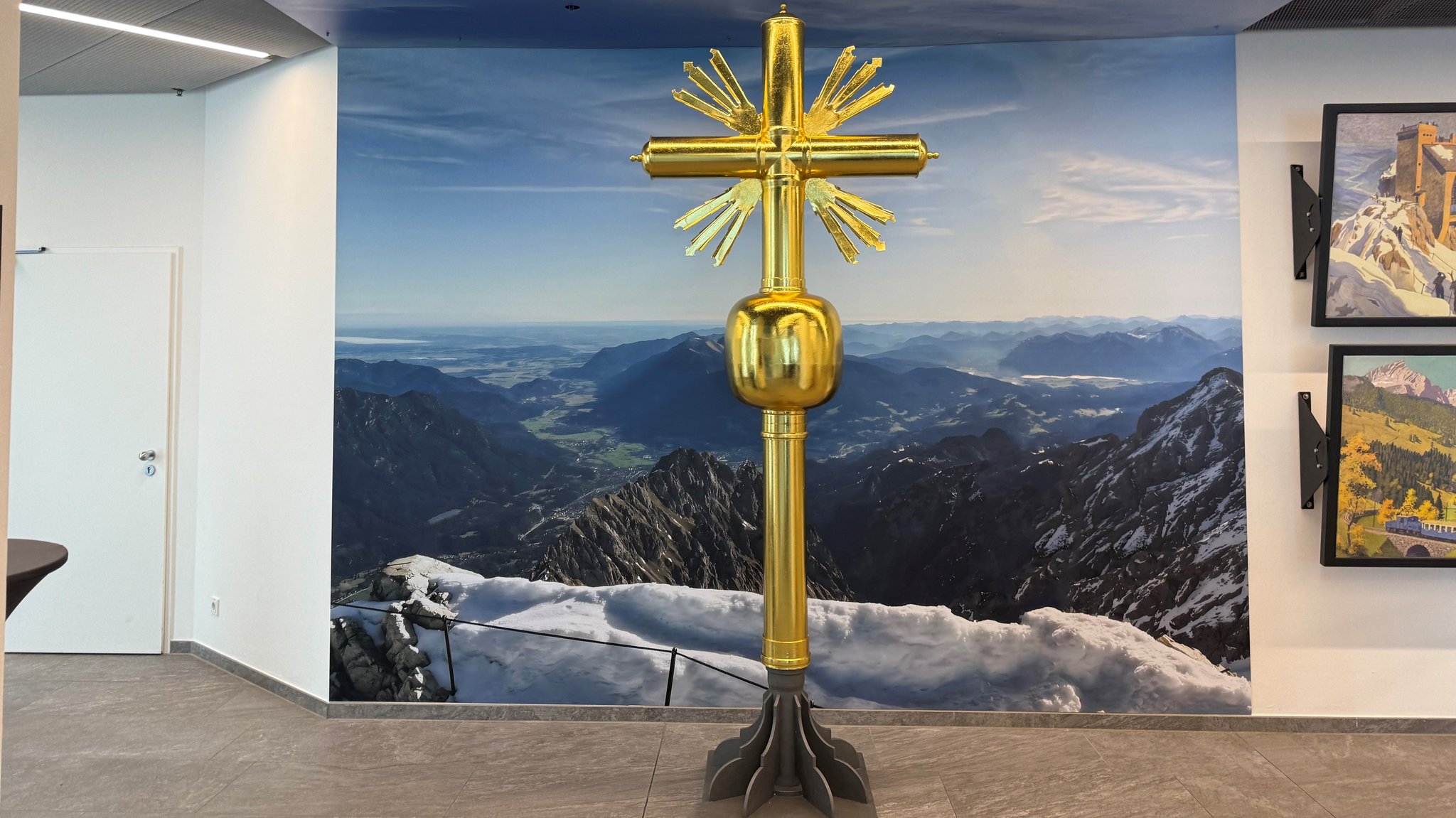 Das neue Zugspitzkreuz ist, wie das Original ebenfalls vergoldet. Es steht in der Gipfelstation der Zugspitzbahn vor einer Fototapete mit Bergpanorama.