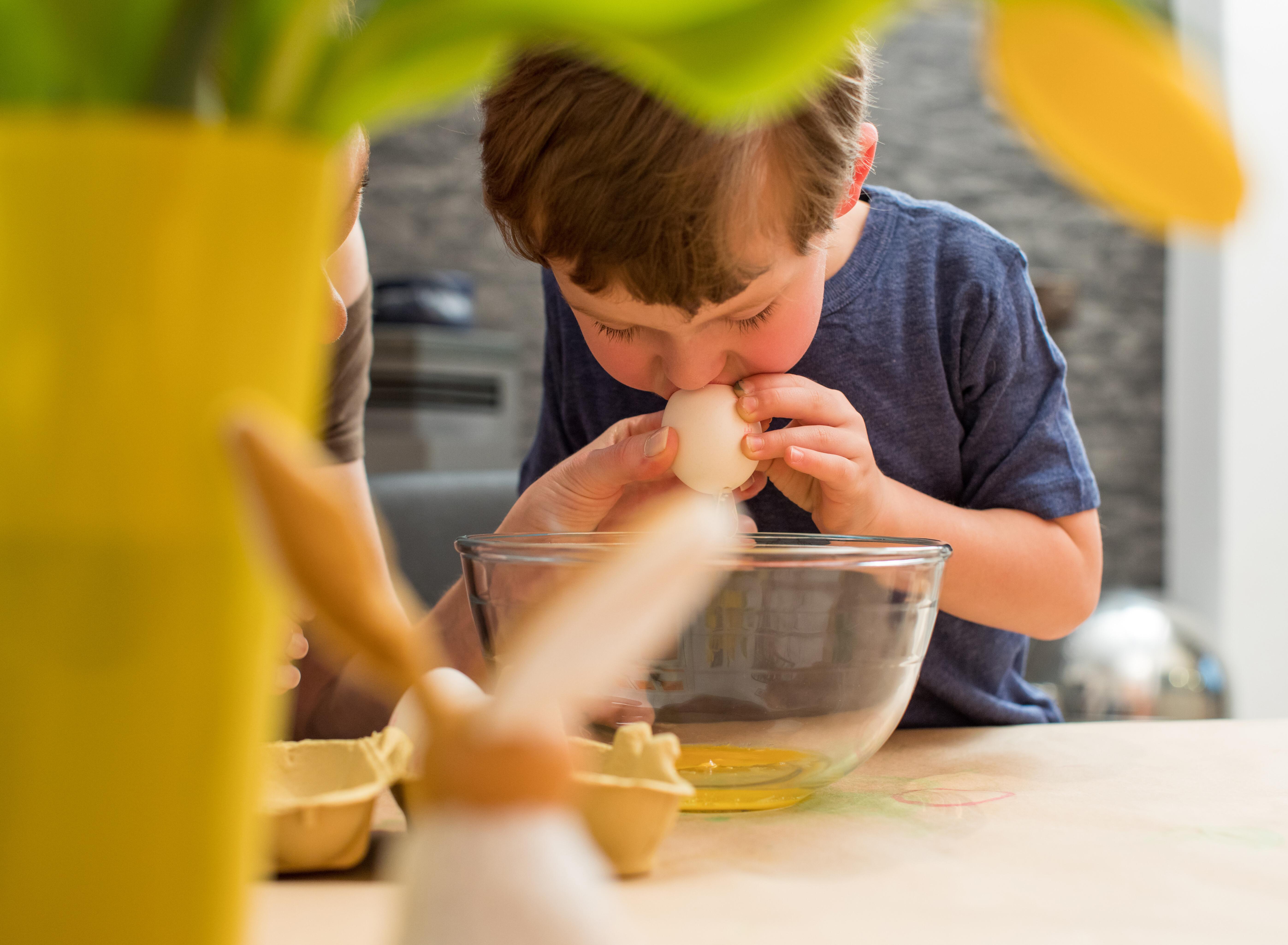 Ein kleiner Junge bläst ein Hühnerei aus. Daneben österliche Dekoration