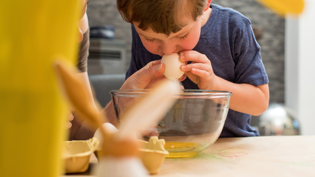 Ein kleiner Junge bläst ein Hühnerei aus. Daneben österliche Dekoration