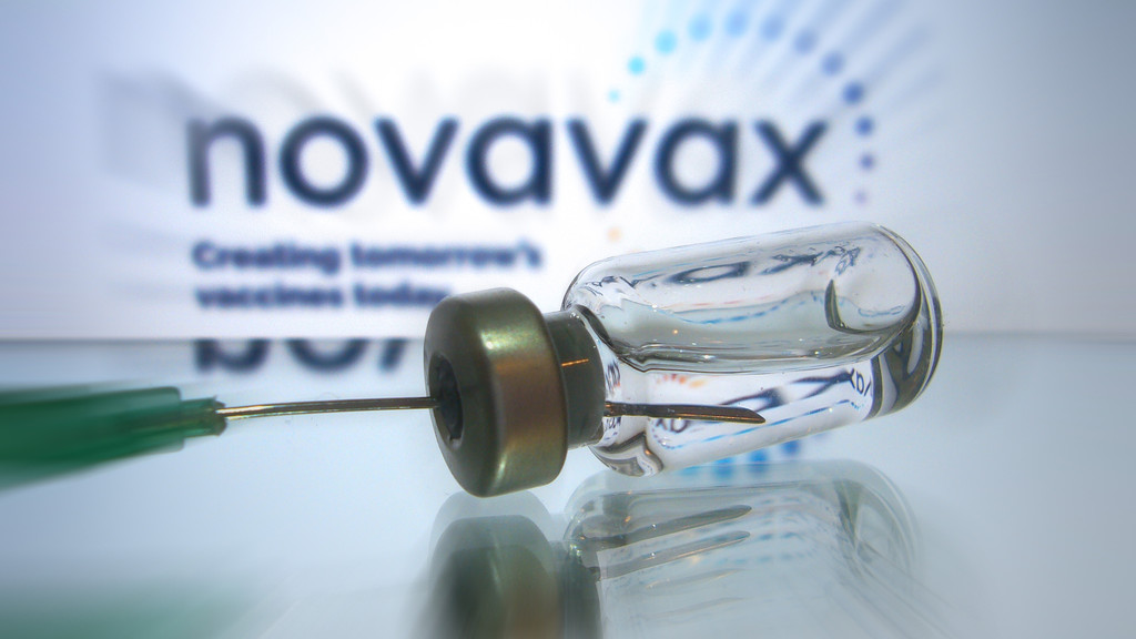 Der Impfstoff von Novavax sollte eigentlich möglichst viele Menschen von einer Impfung überzeugen. Jetzt gerät das Vakzin wegen möglicher Nebenwirkungen in Verruf. Im Bild: Impfdose mit Impfstoff zur Injektion mit einer Kanüle. Im Hintergrund: Schriftzug des Unternehmens Novavax.