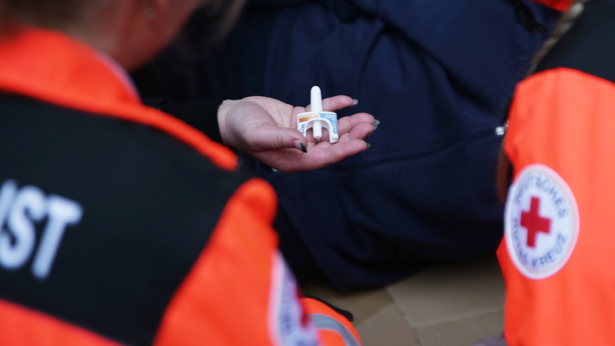 Ein Mann liegt ab Boden, eine Frau hält ein Naloxon-Nasenspray in der Hand. 