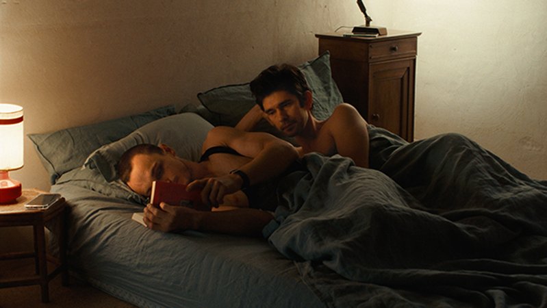 Zwei Männer liegen mit nackten Oberkörpern nachts im Bett. Einer liest, der andere betrachtet seinen Rücken.   